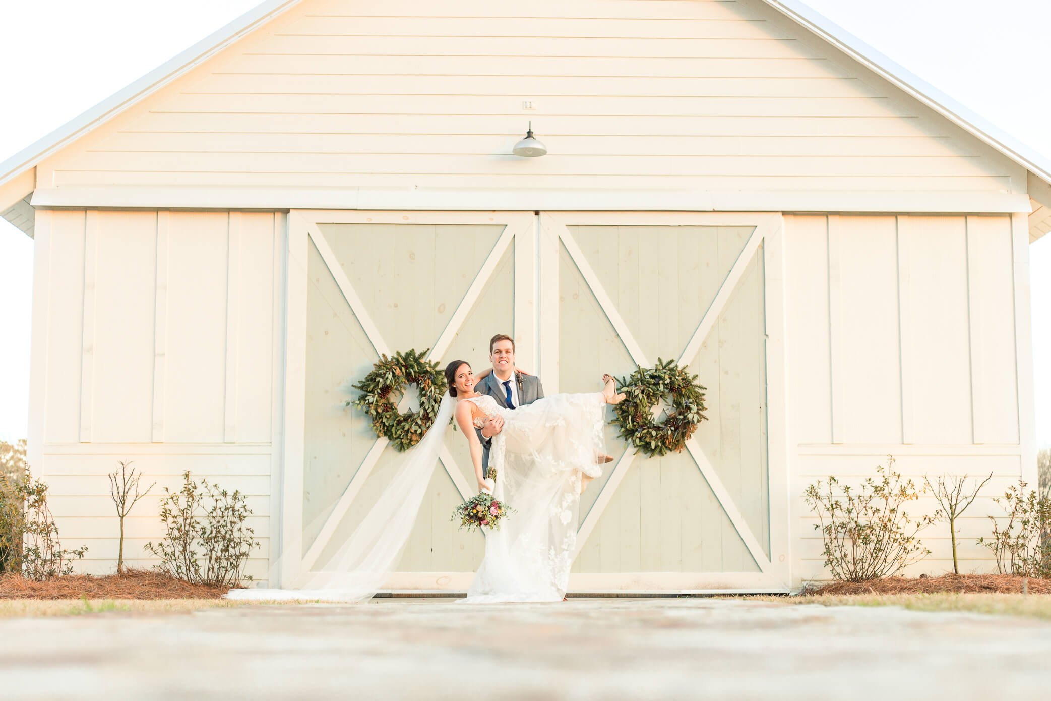 A groom holds and cradles his bride in front of a barn wedding venue in Oxfordshire, as she pops her leg out and holds her bouquet.