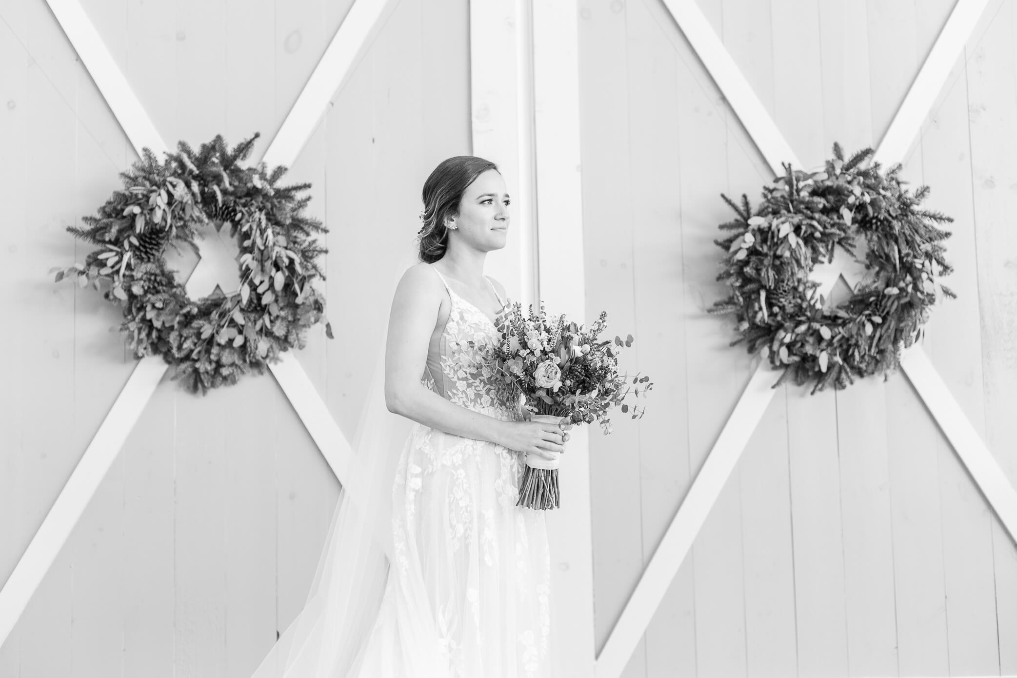 Black and white image of a beautiful bride standing in front of barn doors and holding her bouquet before walking down the aisle.