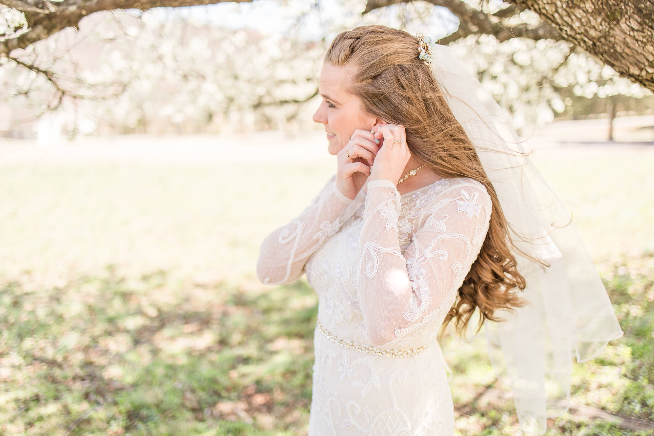 A bride stands beneath a cherry tree in spring as she fixes her earring.