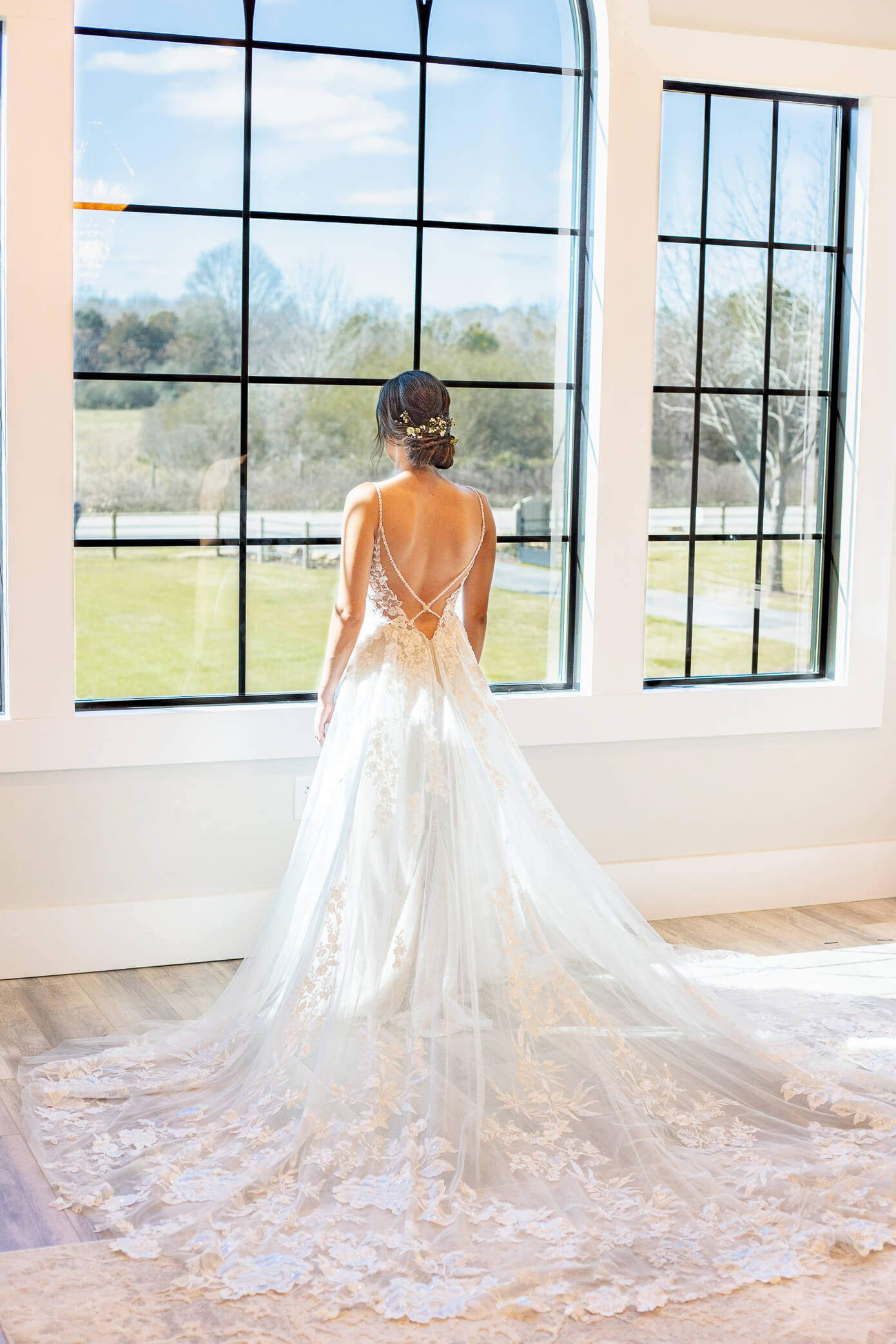 A bride stands looking out a tall window with her dress laid out beautifully in the back. Her updo by a hair salon in Oxford is on display.