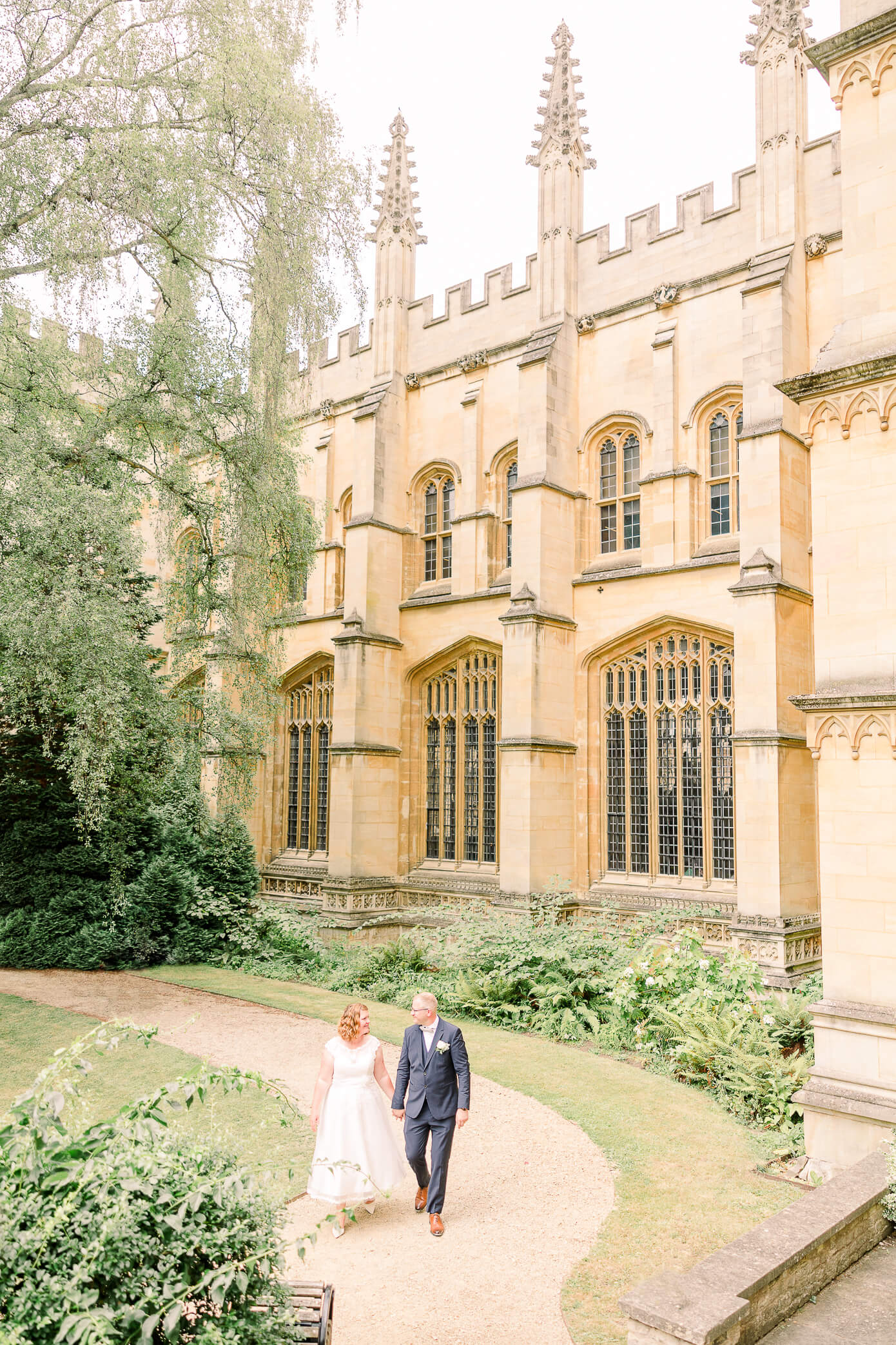 Newlyweds holding hands and walking together along a pathway with a beautiful historic building in the background.