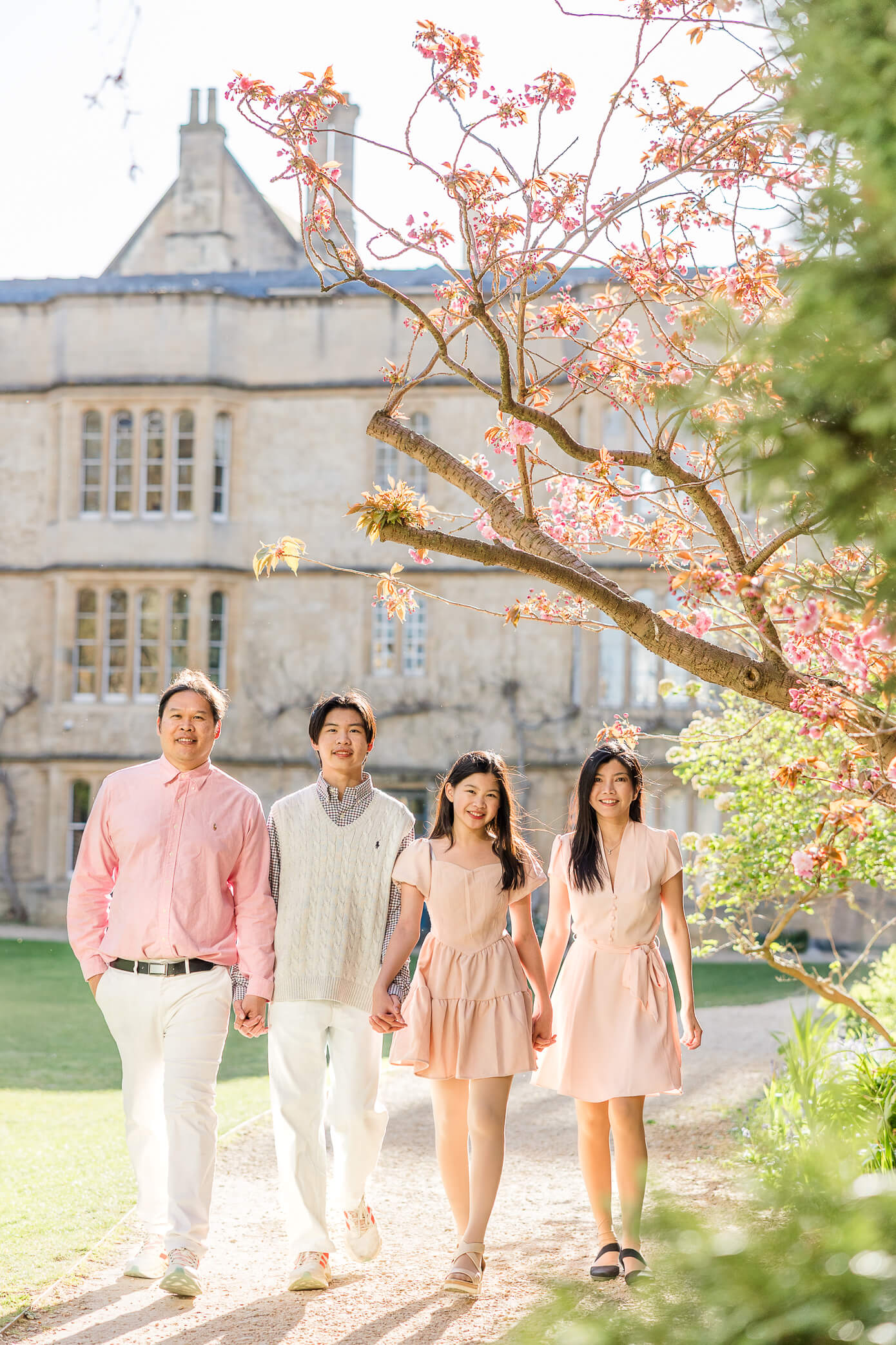 A family with two teenage children pose together holding hands beneath a beautiful pink blossoming tree and an Oxford college building behind.
