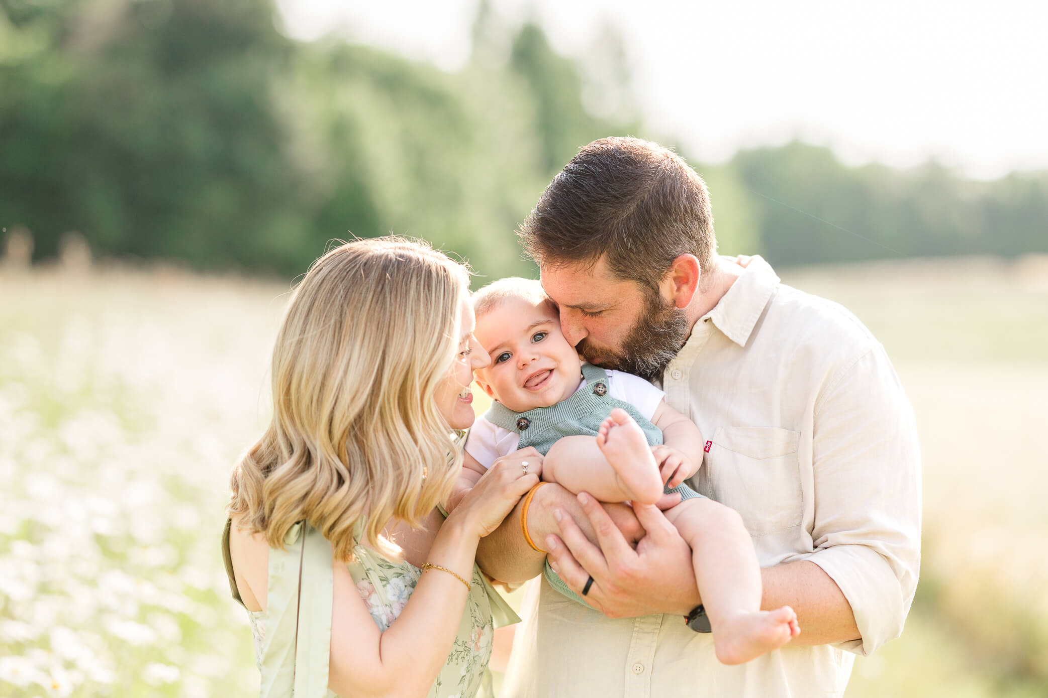 A mother and father both kiss their baby boy's face at the same time as he giggles and smiles and kicks his feet up.