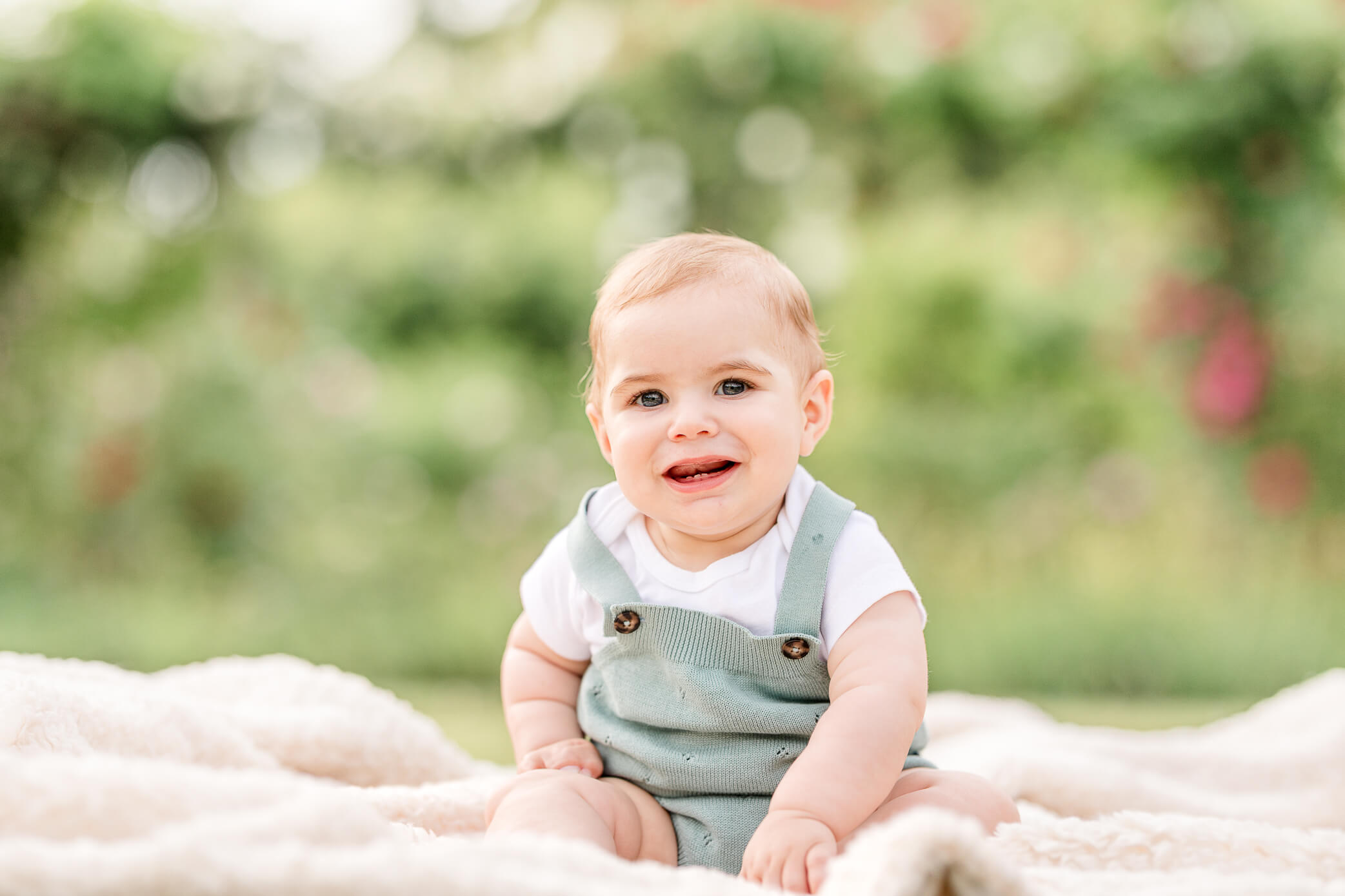A baby sits on a blanket in some grass and smiles a big beautiful smile while his parents find things to do in Oxford with kids.
