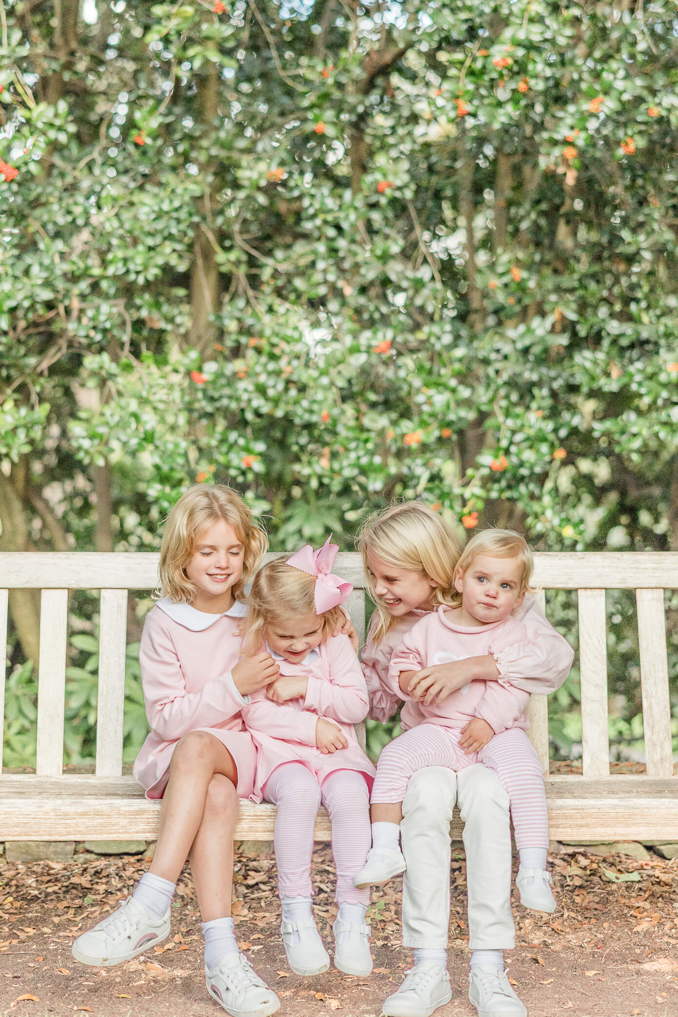 Four young, blonde girls who are sisters sit together on a bench in front of a big hedge. They tickle each other and laugh together.