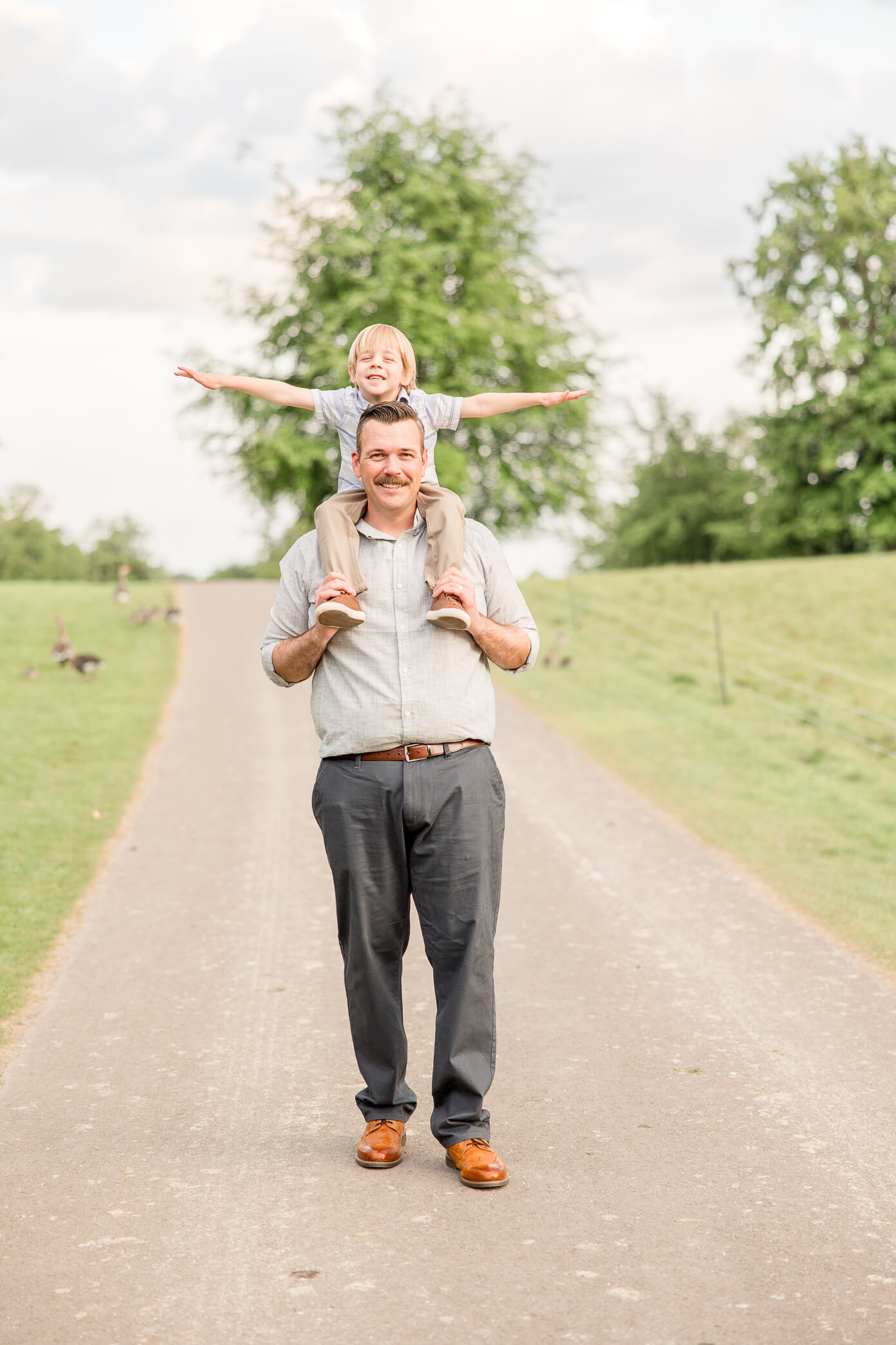 a little boy riding on his dad's shoulders and holding his arms out like an airplane while walking through a park.
