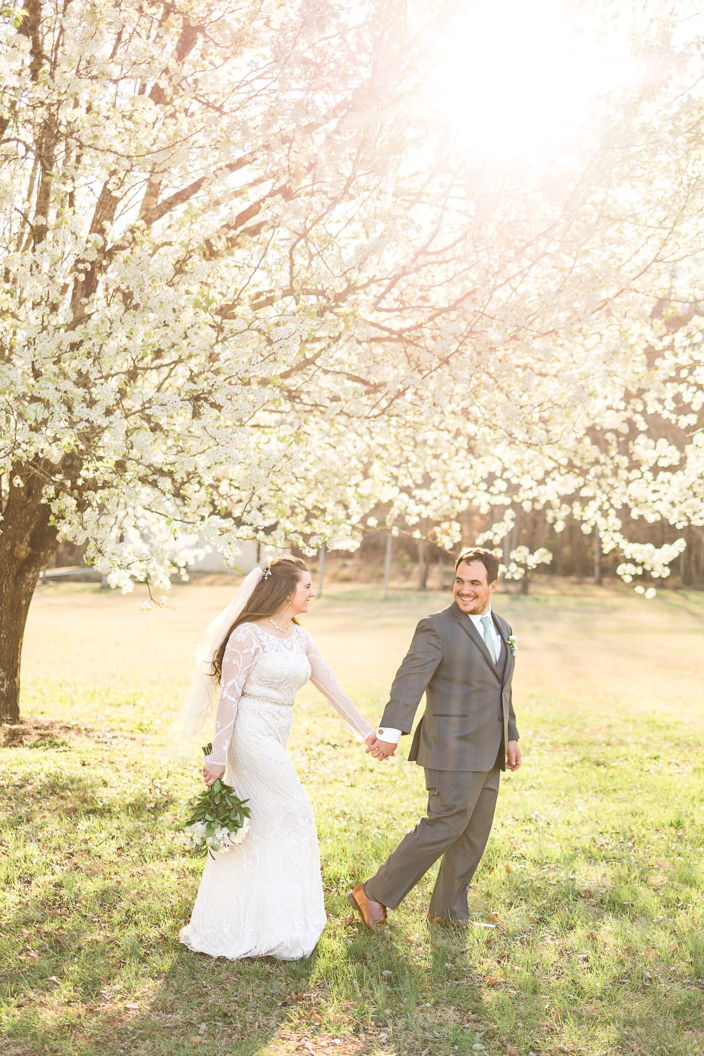 A groom leads his bride by the hand as they walk in front of a gorgeous cherry blossom tree with the sun setting behind