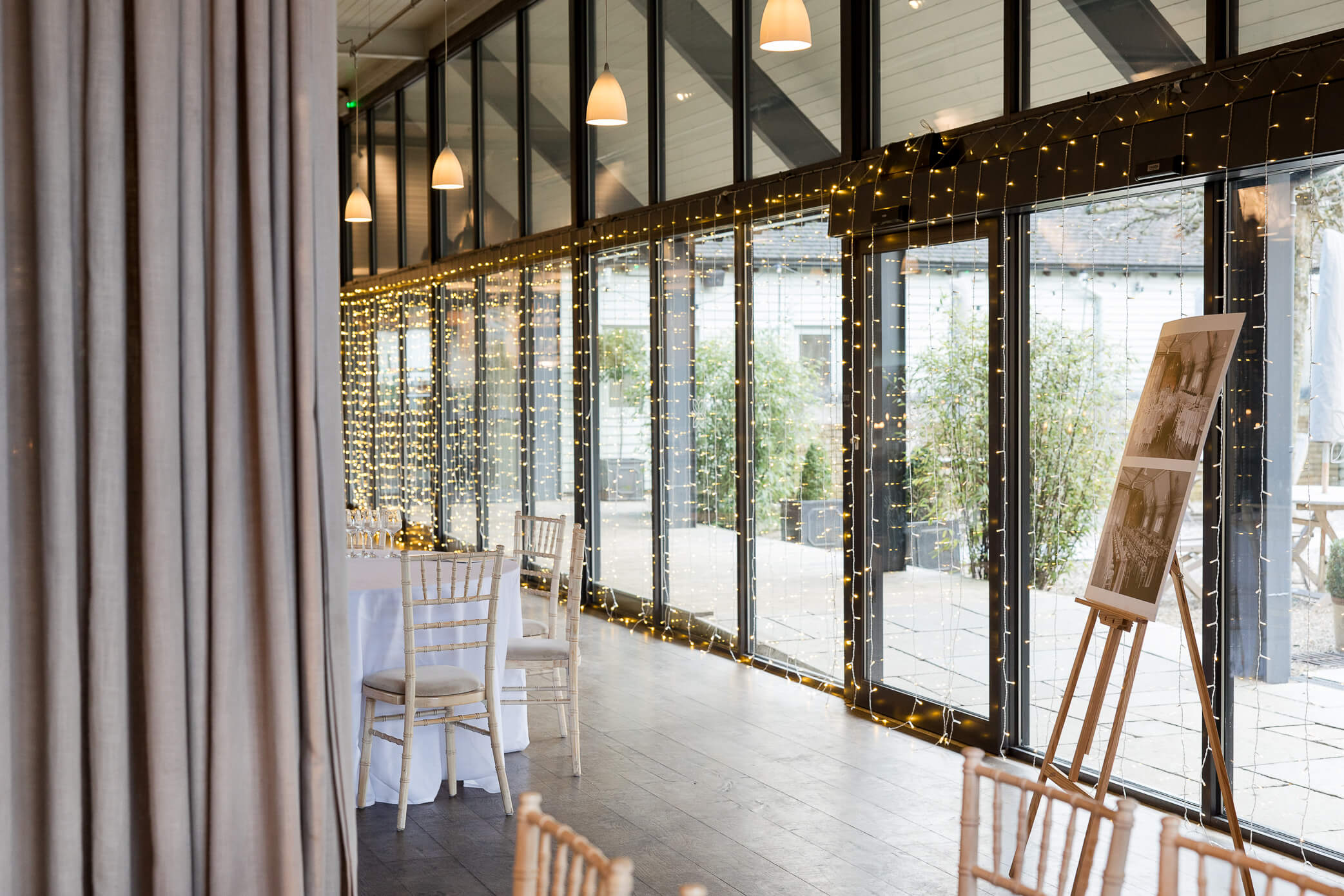 The dining hall and reception room at Lapstone Barn wedding venue, showcasing fairy lights waterfalling from the large wall of windows.