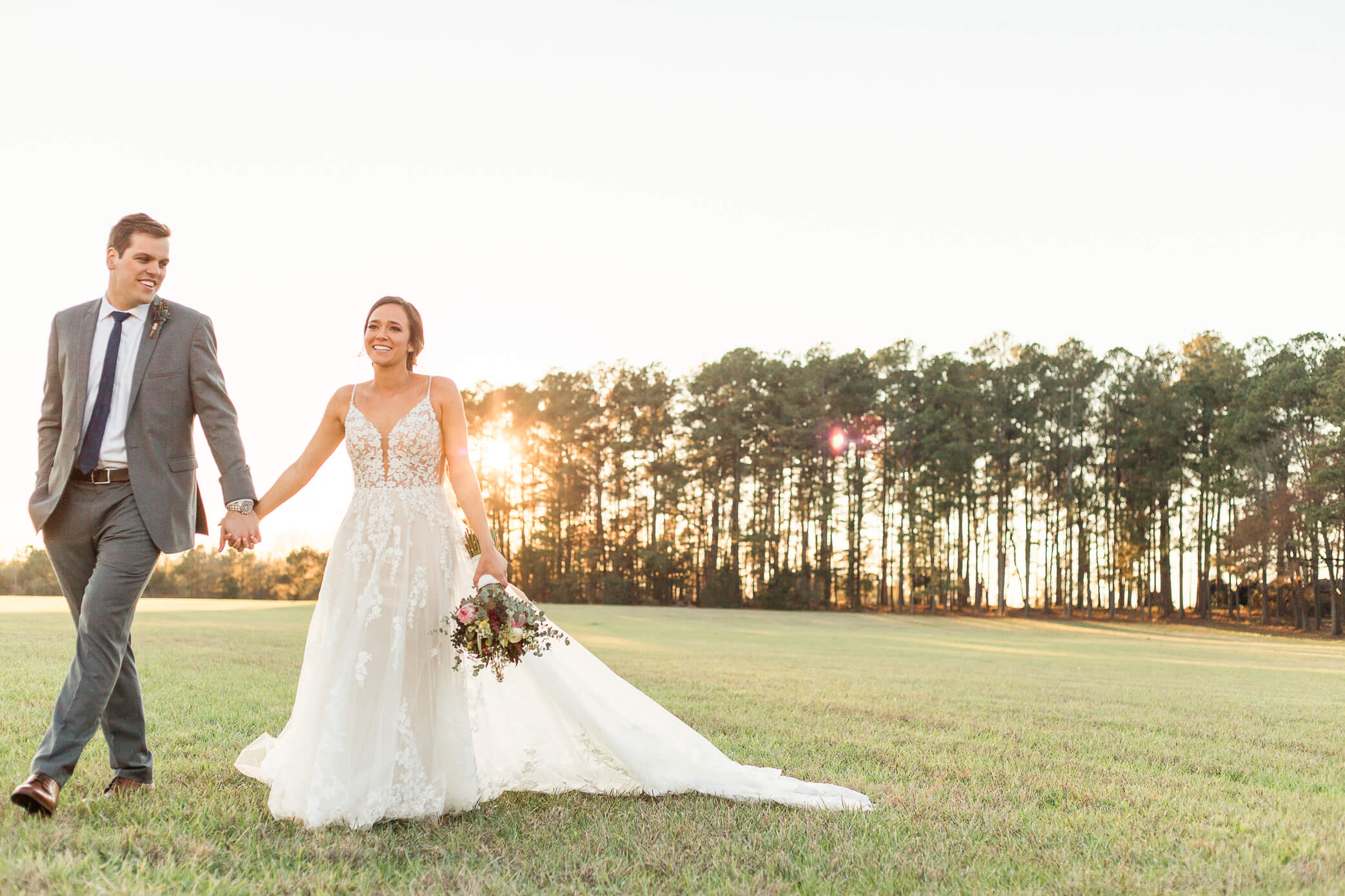 A bride and groom walking hand in hand at golden hour in the countryside.