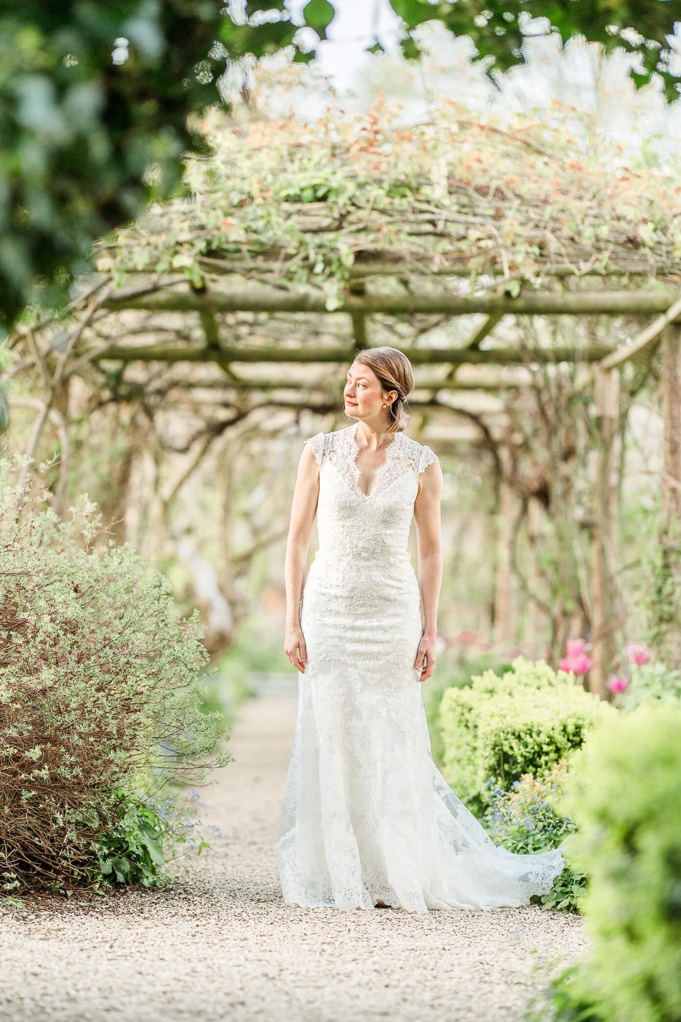A bride stands on a gravel path at a Rousham House wedding as she looks down the garden pathway with a pergola behind her.