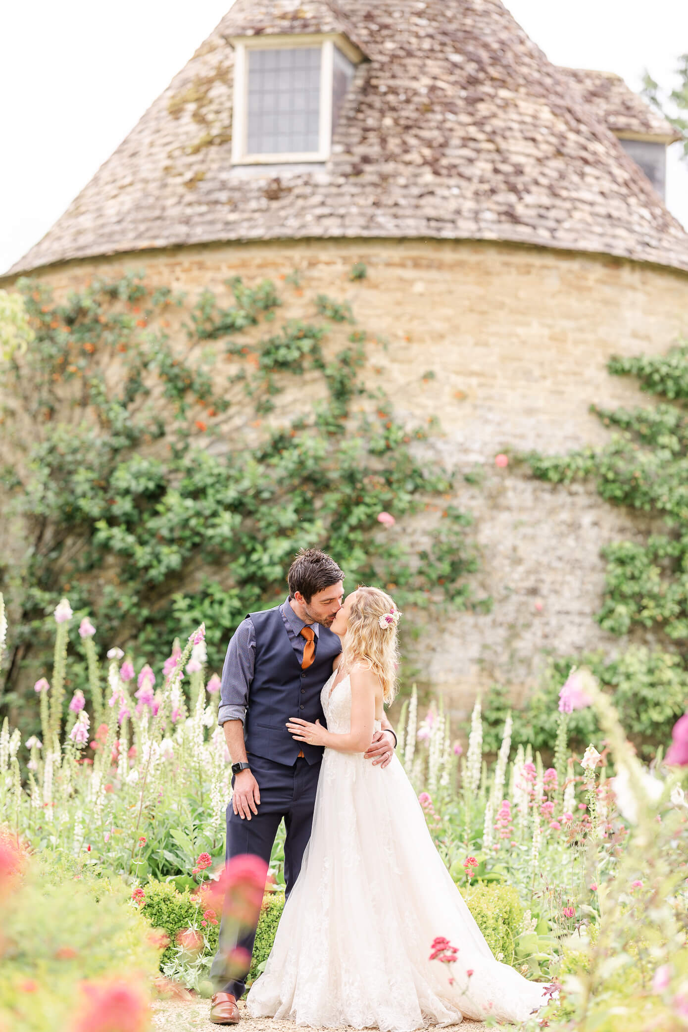 A groom and bride kissing in front of a dovecote garden and surrounded by foxglove flowers.
