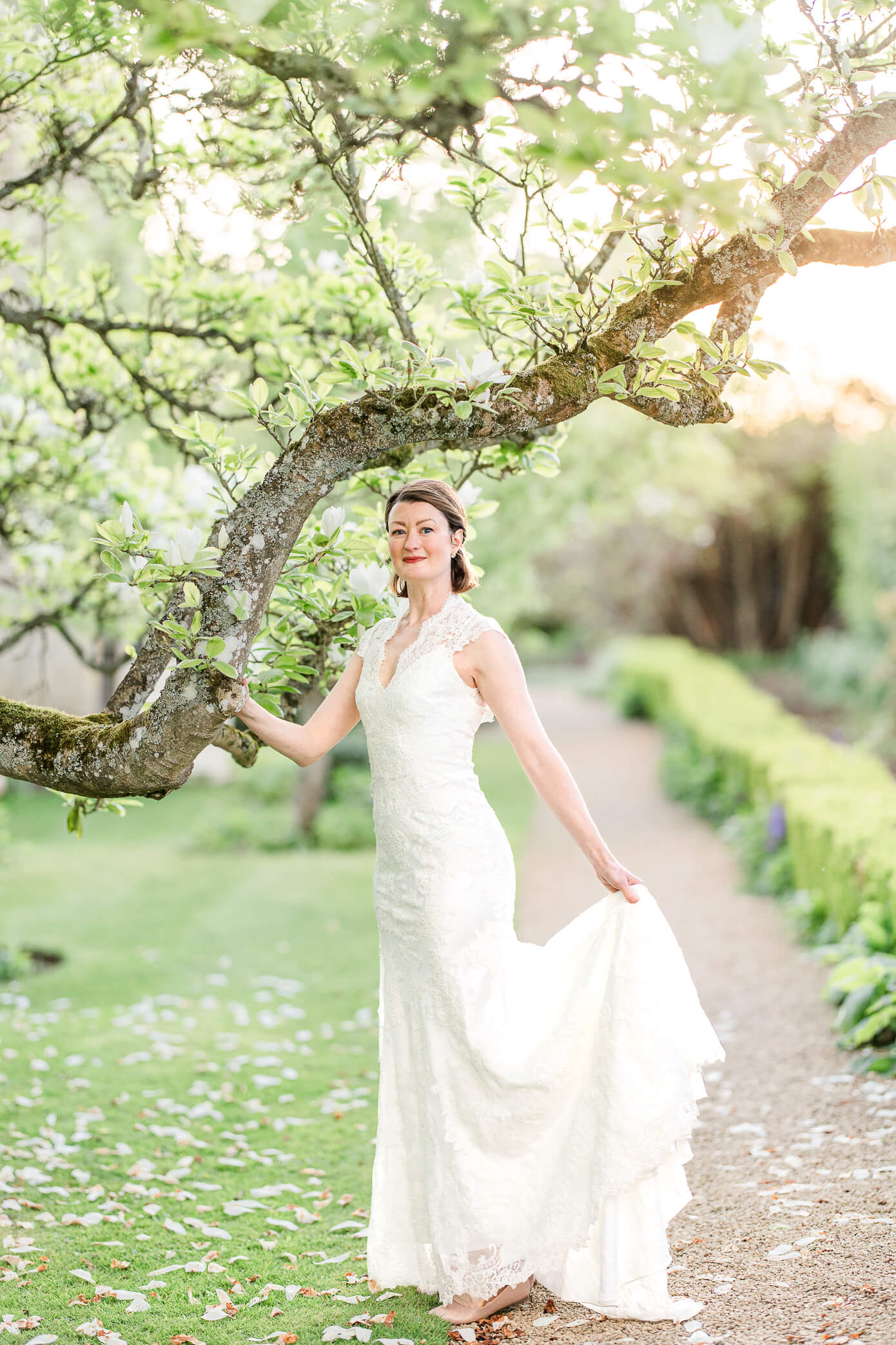 A bride poses by a low twisting branch and holds her dress in her left hand as the sun is setting behind at a Rousham House wedding.