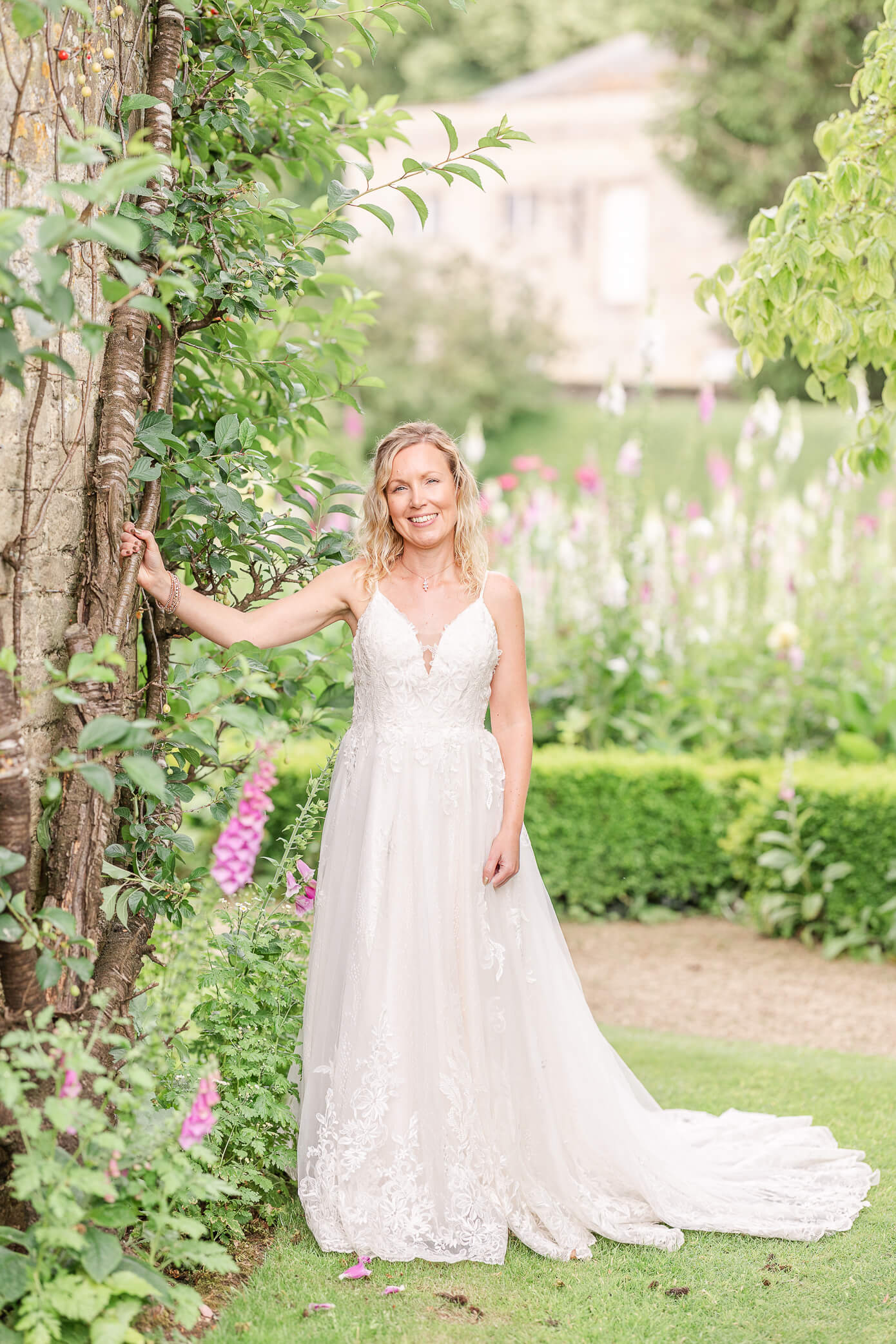 A bride poses next to an old dovecote in a garden. She rests her hand on the building and smiles.