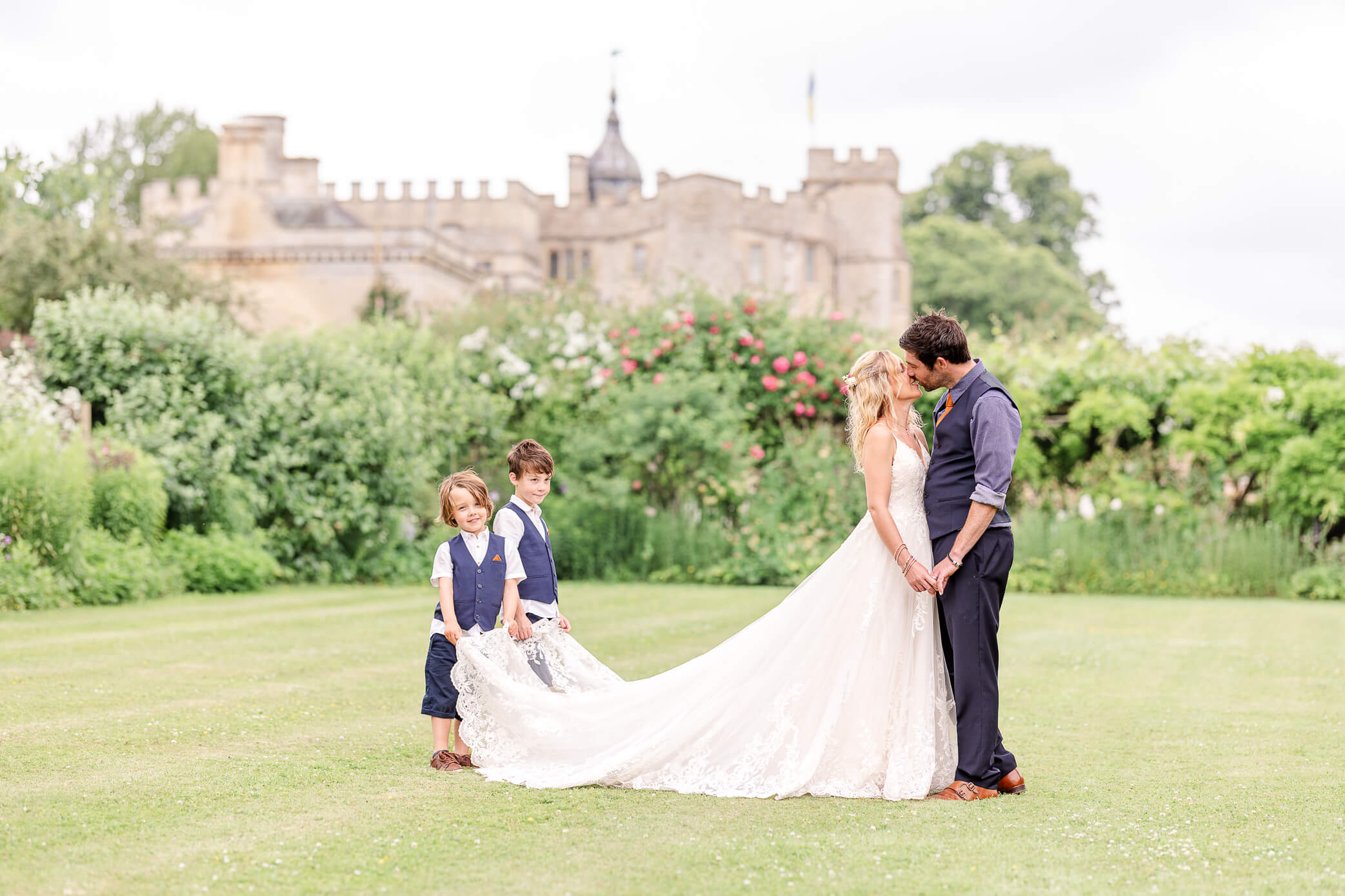 Bride and groom kiss while their two sons hold her dress up from the ground. There is a beautiful manor house in the background.