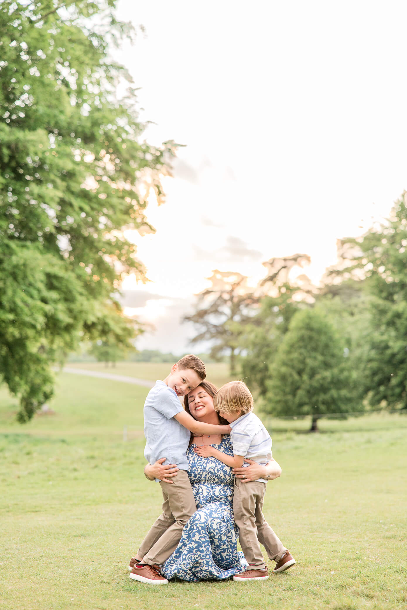 Two boys hug their mother really big as she kneels down in the grass on their level