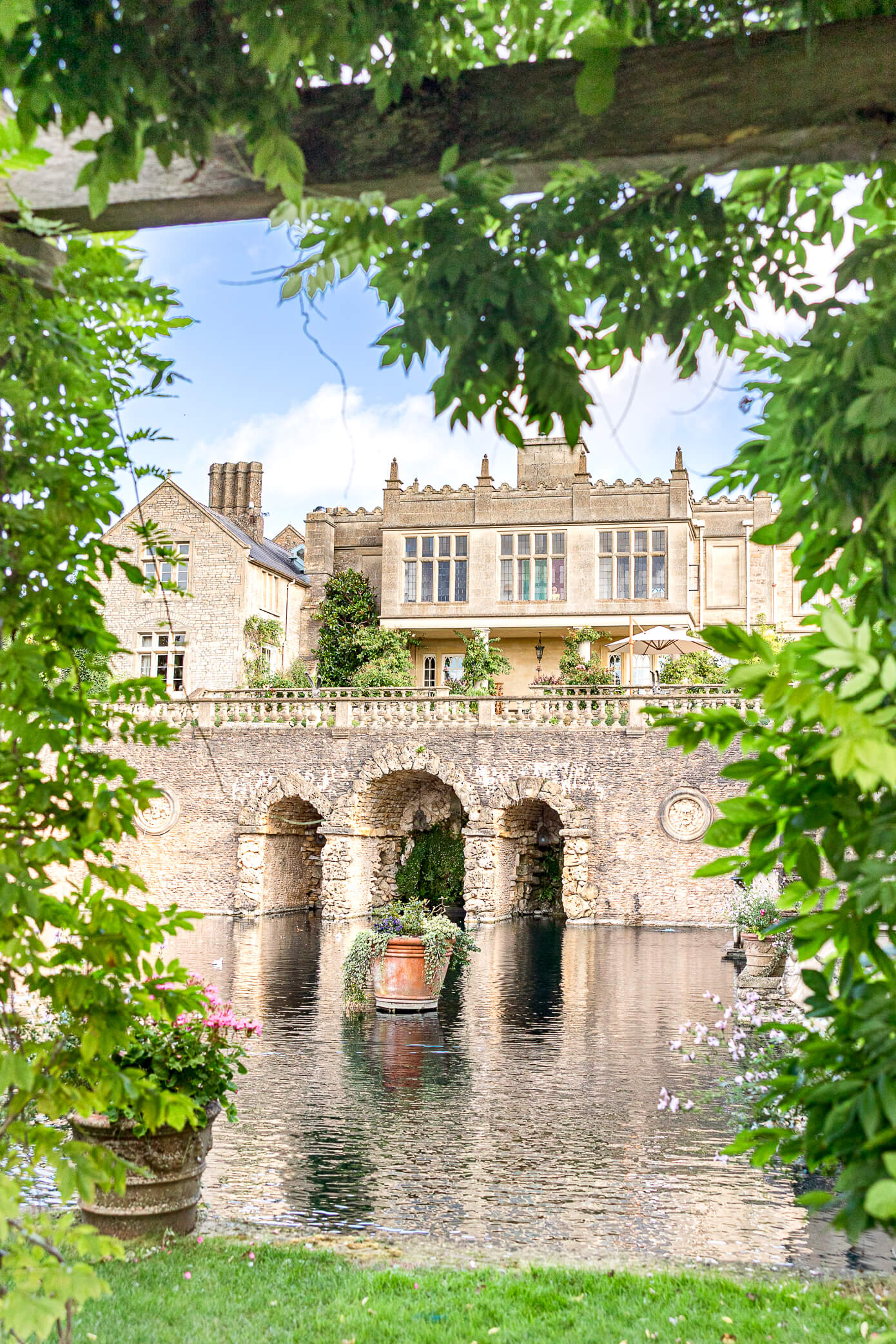 A view of Euridge Manor's stone arched bridge from beneath a pergola covered in greenery