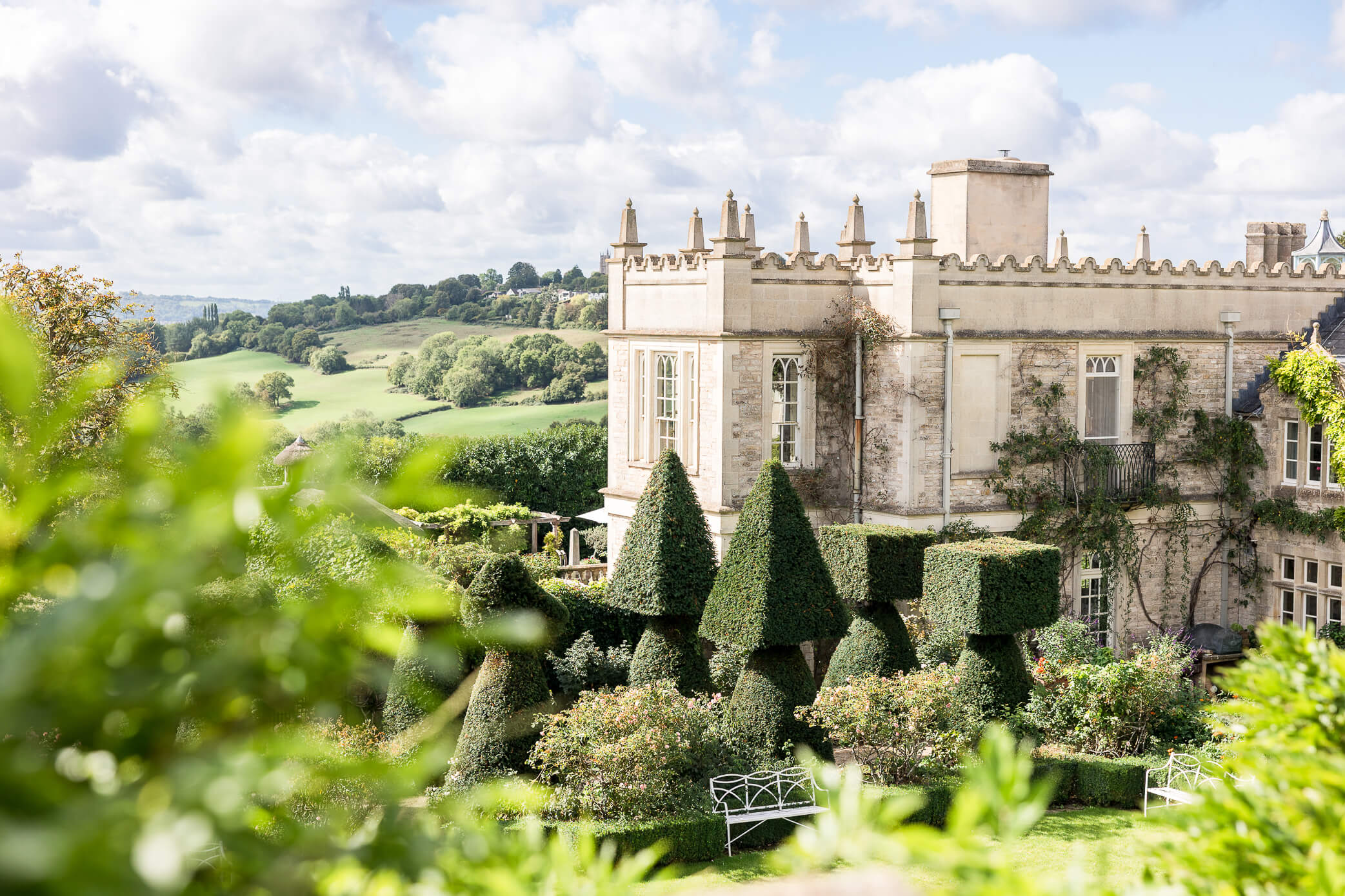 The view of the main house at Euridge Manor from atop the Orangery Terrace, through some greenery, and beneath a beautiful sky